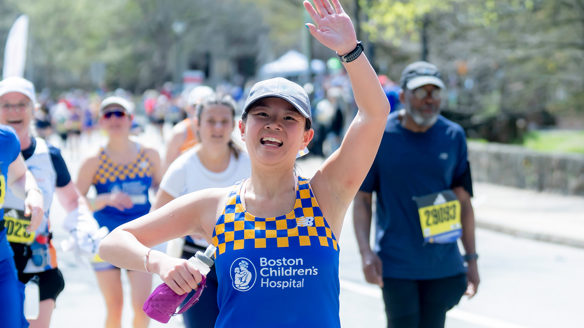 A person wearing a navy bule baseball cap and bright blue and yellow check Boston Children's Hospital singlet runs with her arm raised waving.