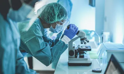 A group of researchers wearing personal protective equipment stand at a table, and one looks into a microscope