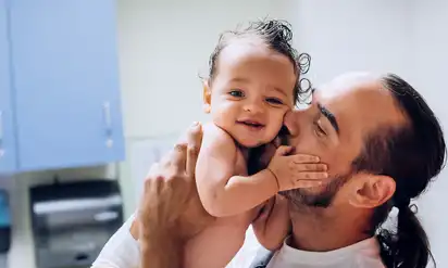 A smiling baby patient being held and kissed by his father in a hospital room