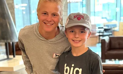 Two smiling boys pose together indoors; the older teen in a gray hoodie leans in with an arm around a younger boy wearing a gray cap and a dark T-shirt, with seating blurred in the background.