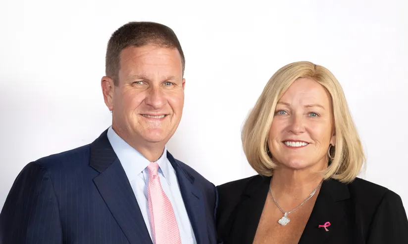 Rob Hale (a man with dark, close cropped hair wearing a suit and a pink tie) stands with his wife, Karen Hale ( a woman with shoulder length blonde hair wearing a dark jacket and a pink shirt). The two are smiling.