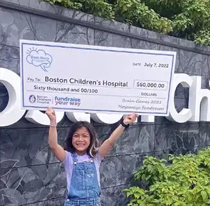 A young girl proudly holds a large check over her head for $60,000 made out to Boston Children's Hospital