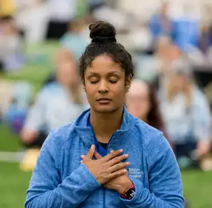 A woman holds her hands over her heart. A number of people sitting on yoga mats can be seen in the background.