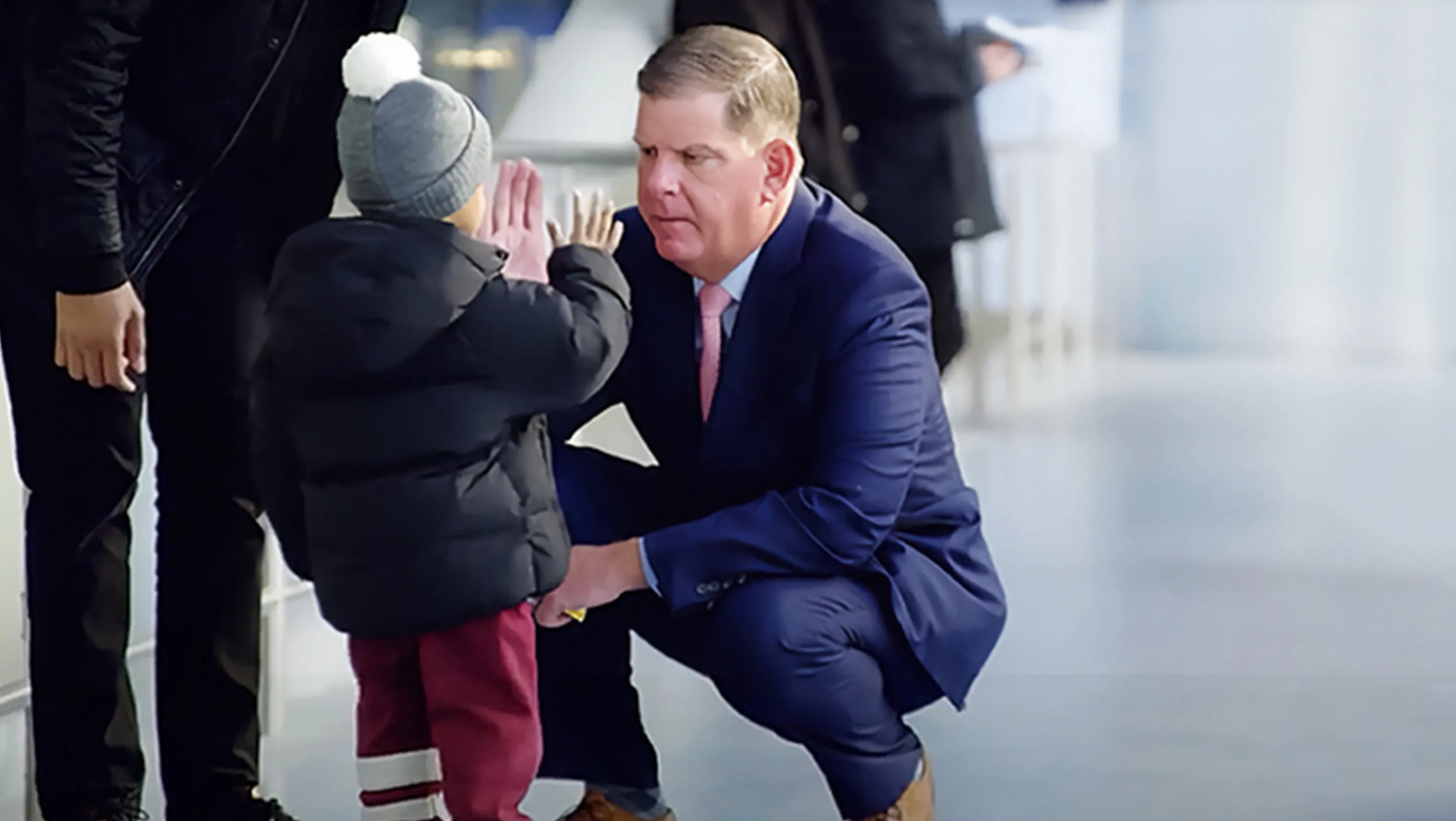 Man in blue suit squatting to high five a child with winter coat and hat