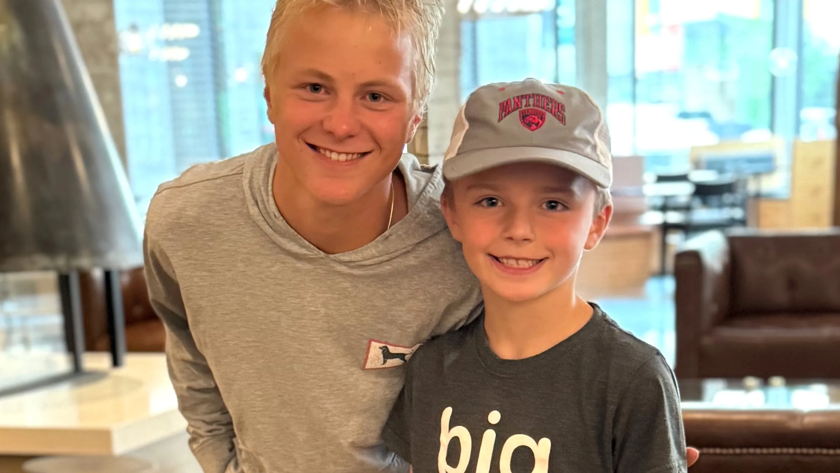 Two smiling boys pose together indoors; the older teen in a gray hoodie leans in with an arm around a younger boy wearing a gray cap and a dark T-shirt, with seating blurred in the background.