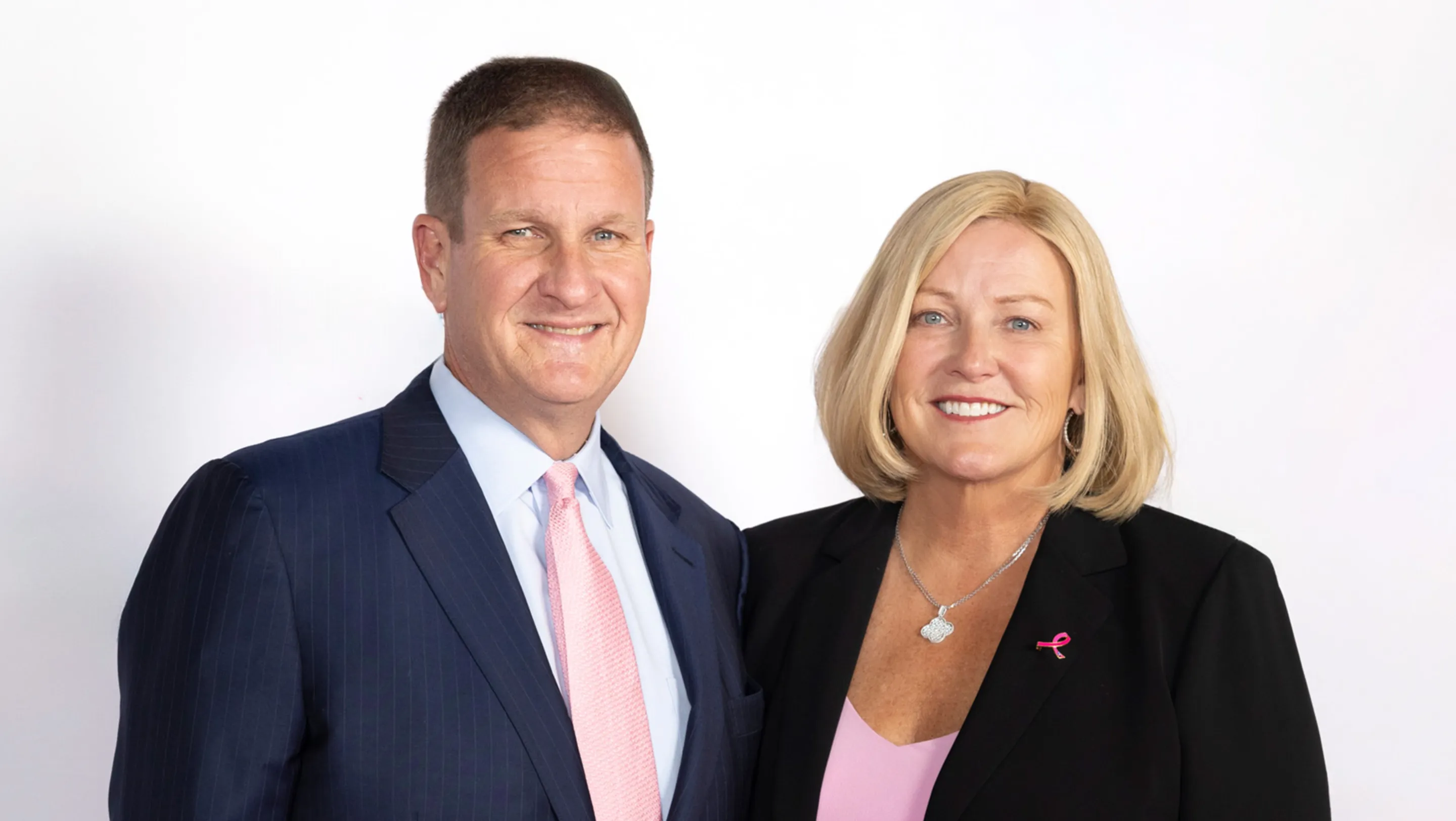 Rob Hale (a man with dark, close cropped hair wearing a suit and a pink tie) stands with his wife, Karen Hale ( a woman with shoulder length blonde hair wearing a dark jacket and a pink shirt). The two are smiling.