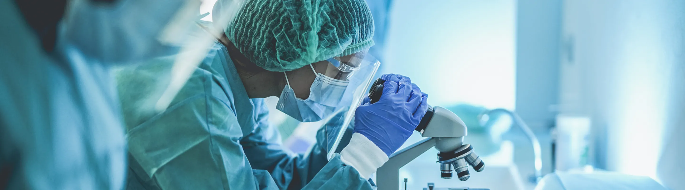 A group of researchers wearing personal protective equipment stand at a table, and one looks into a microscope