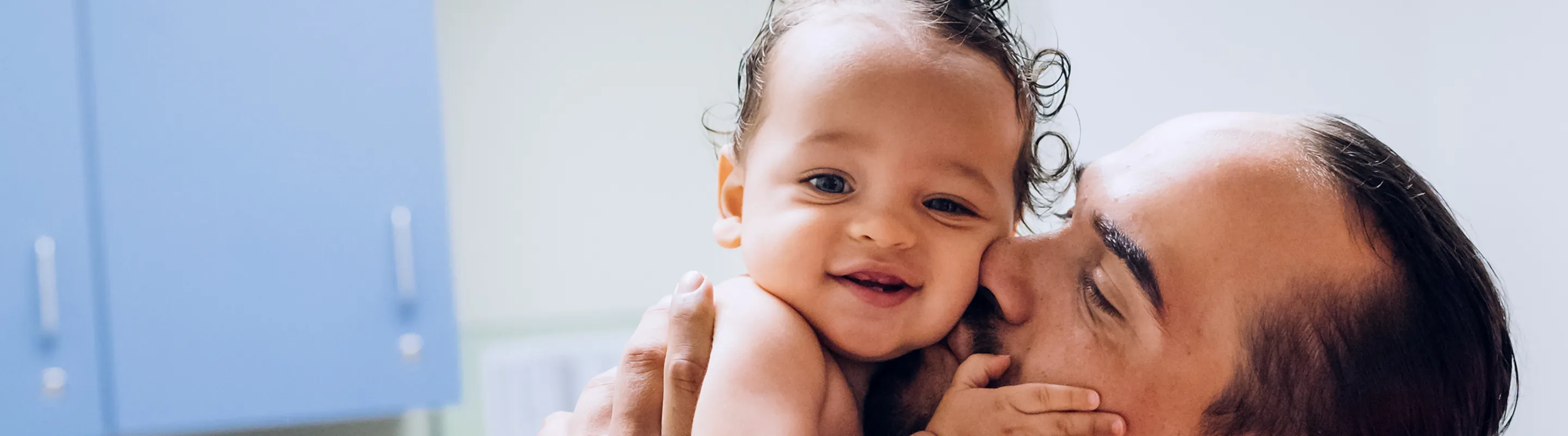 A smiling baby patient being held and kissed by his father in a hospital room