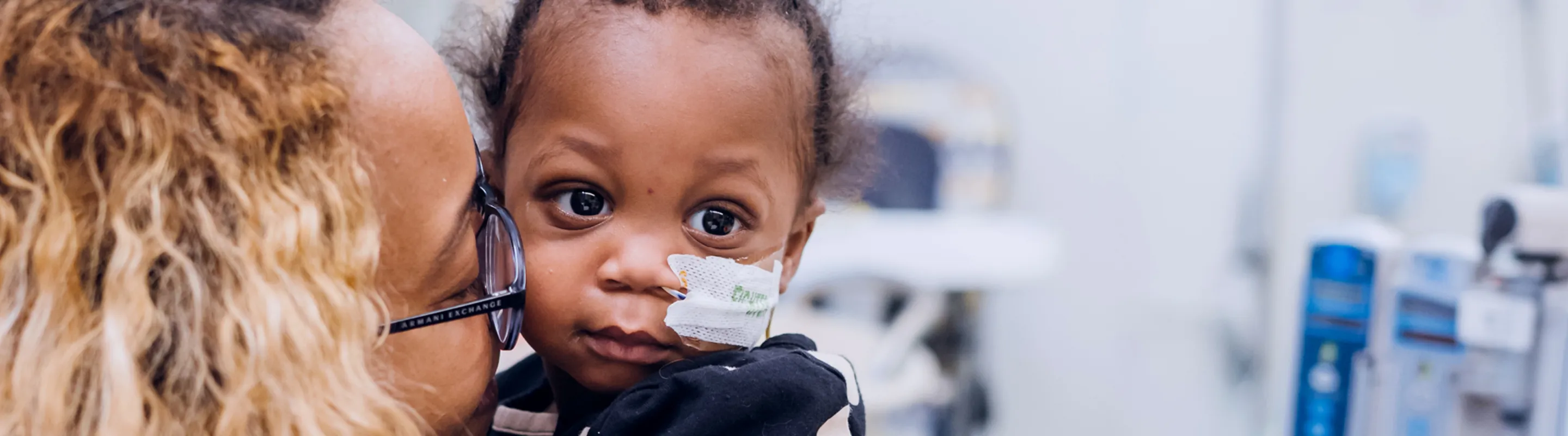 A baby patient with a tube in her nose is held and kissed by her mom, with hospital equipment in the background