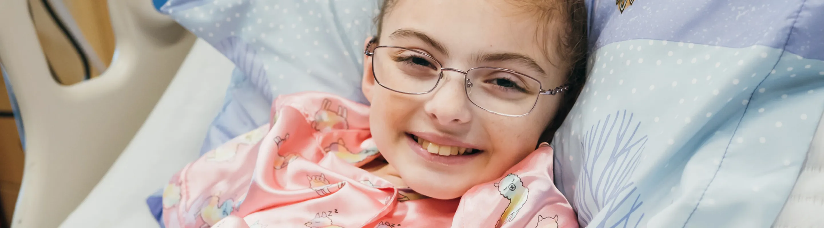 A young girl wearing glasses sits in a hospital bed and smiles at the camera