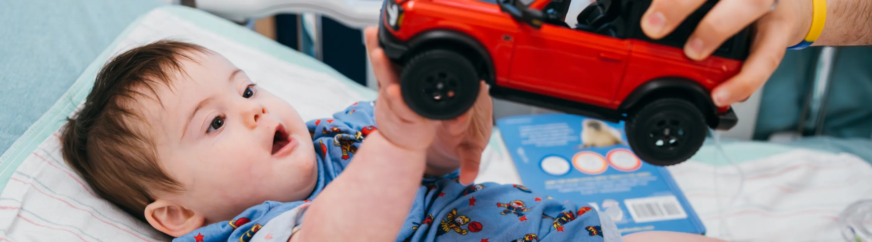 small child lying in a hospital bed grasping at a red toy jeep truck