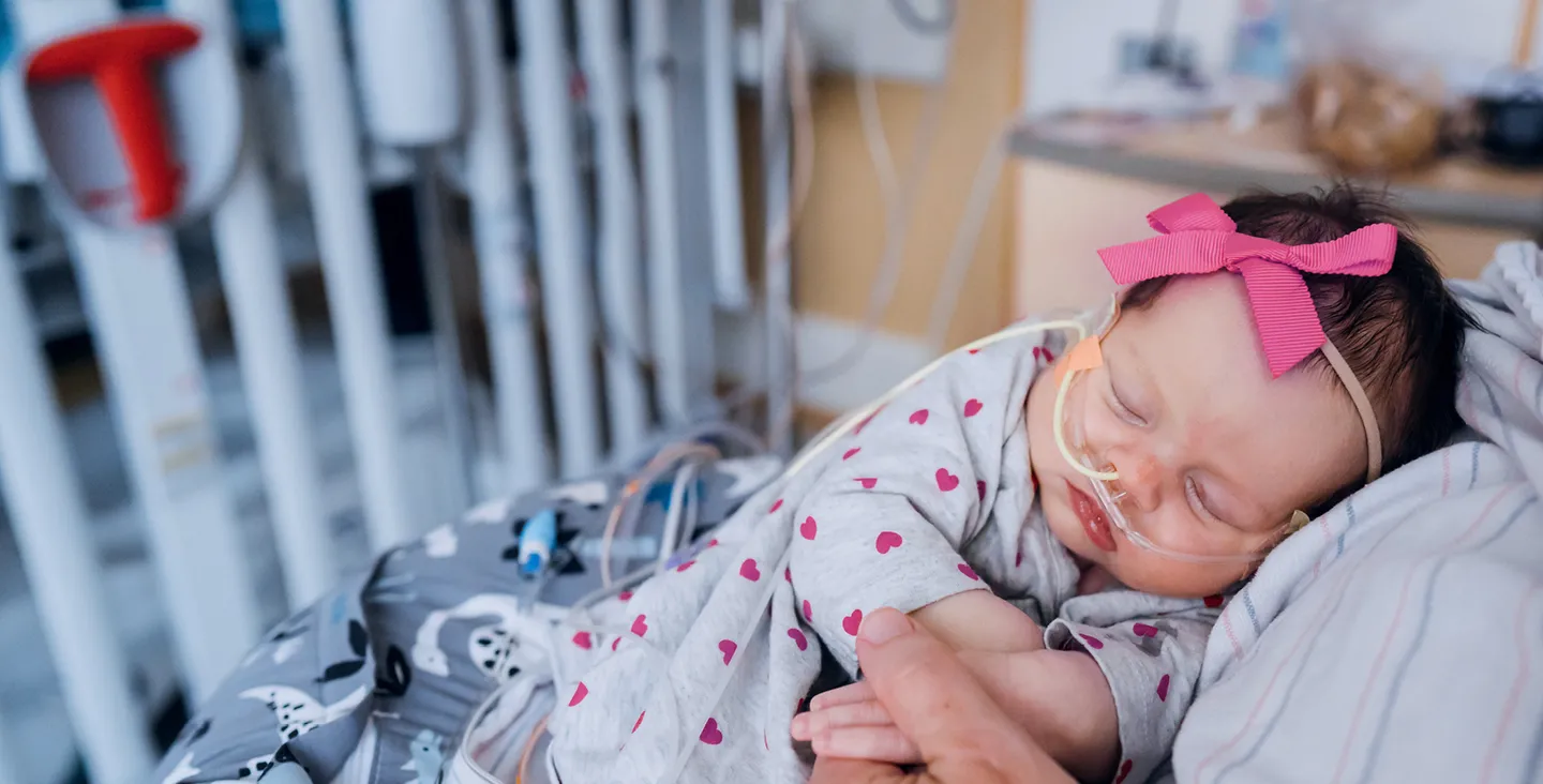 A tiny baby wearing a pink bow with an oxygen tube in her nose is held on her mother's chest in a hospital room