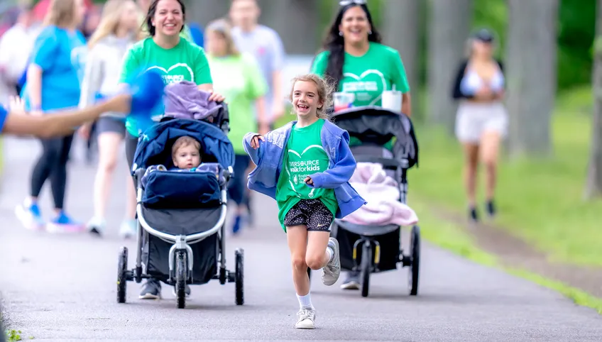 Two smiling women push strollers as a young girl runs ahead of them gleefully
