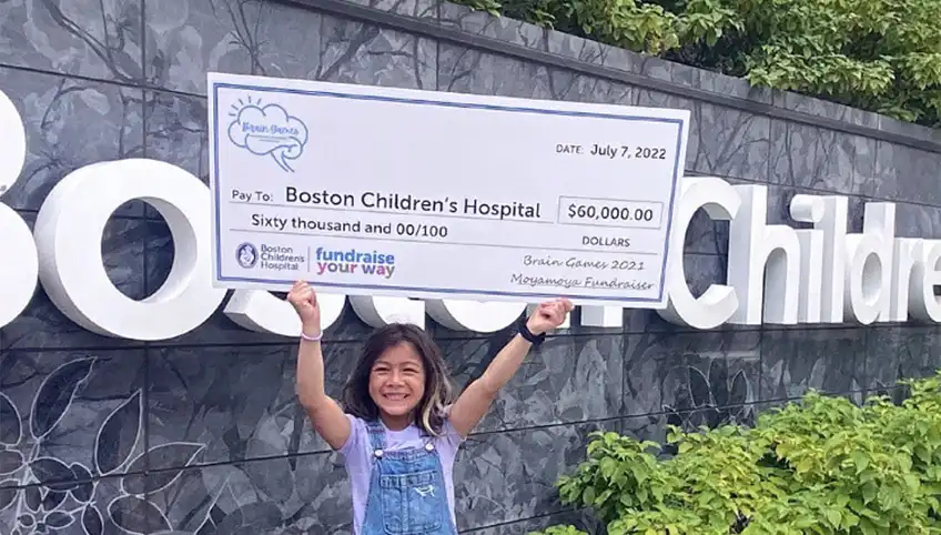 A young girl proudly holds a large check over her head for $60,000 made out to Boston Children's Hospital