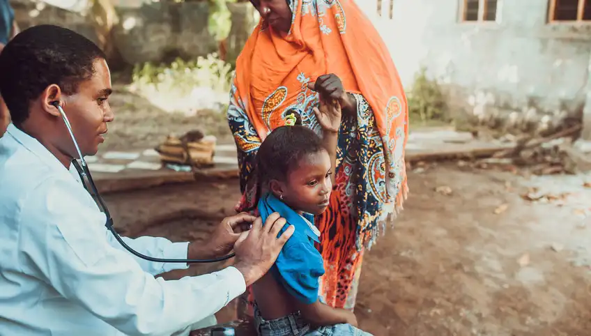 A doctor wearing a stethoscope listens to the heart of a young girl. They appear to be in Africa