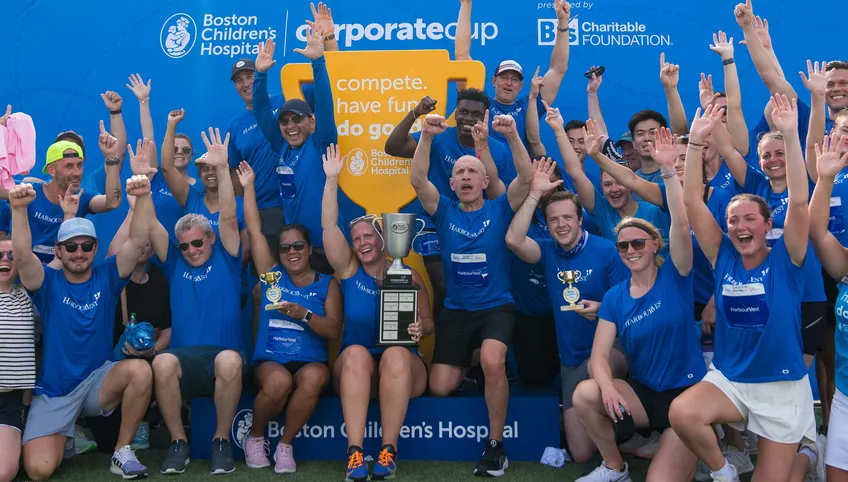 A group of Corporate Cup event attendees wearing matching blue shirts cheer at the awards ceremony. A person in the middle holds a large trophy.