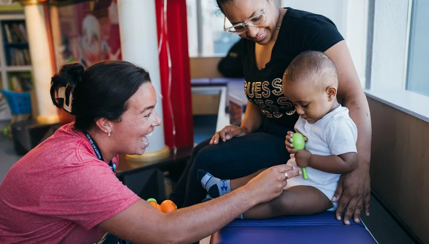 A small baby sitting next to his mom holds a rattle while a nurse smiles and tickles him