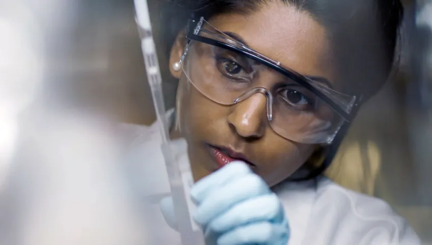 A woman wearing safety glasses and gloves examines a test tube