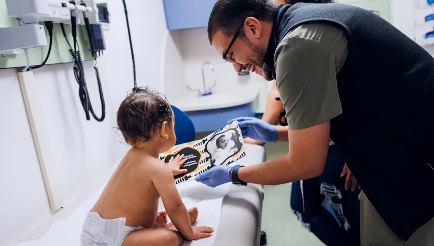 A baby patient sitting on a hospital exam table looks at a card being held open to him by his father
