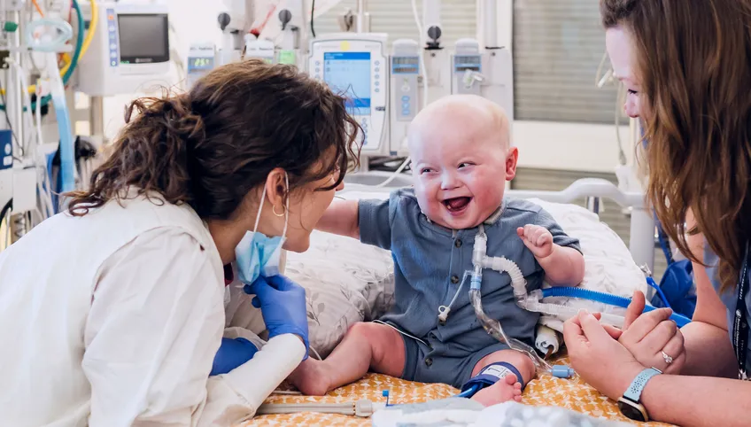 A baby patient surrounded by medical equipment site on a hospital bed laughing while a doctor and his mom smile back