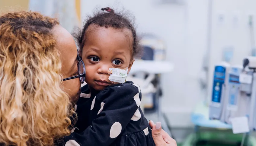 A baby patient with a tube in her nose is held and kissed by her mom, with hospital equipment in the background