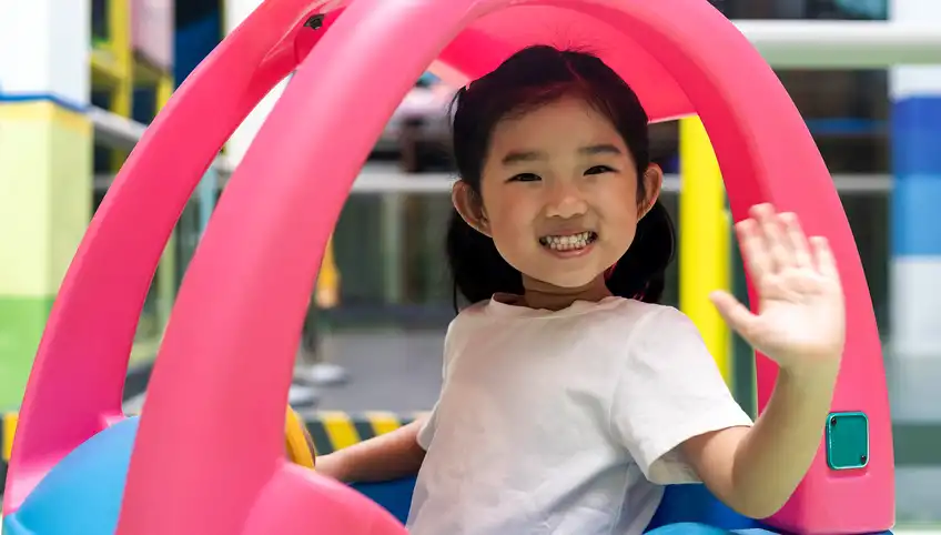 A young girl rides in a toy car and waves at the camera