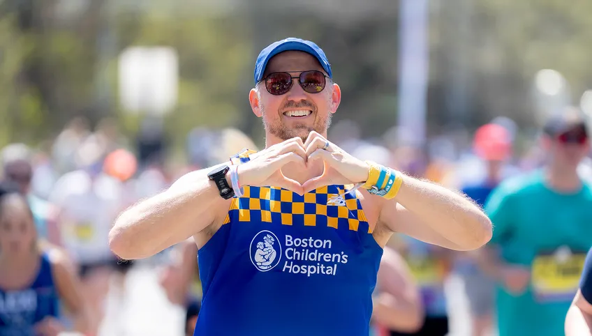 A person wearing a blue baseball cap, sunglasses and a blue Boston Children's Hospital running single with yellow checks makes a heart with their hands