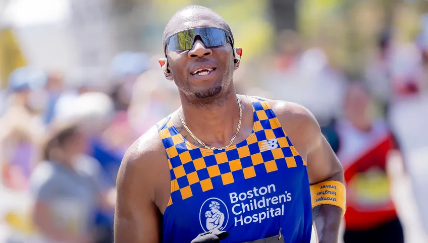A person wearing sunglasses and a blue Boston Children's Hospital running singlet with yellow checks smiles at the camera while running