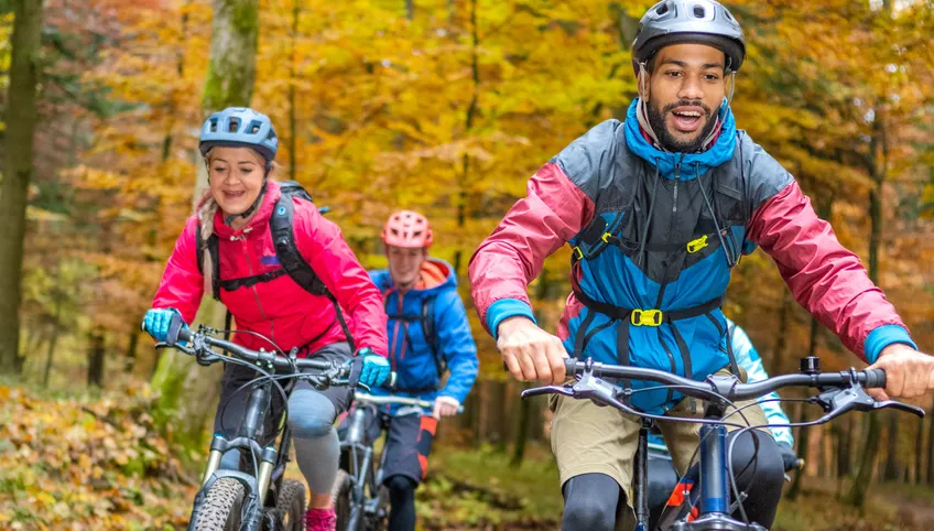 a group of people ride bikes in a wooded area. All wear helmets and are on mountain bikes. 