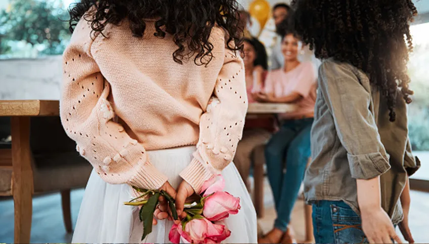 Two people with long, curly hair stand in front of a table with balloons. One person holds pink flowers behind their back. 