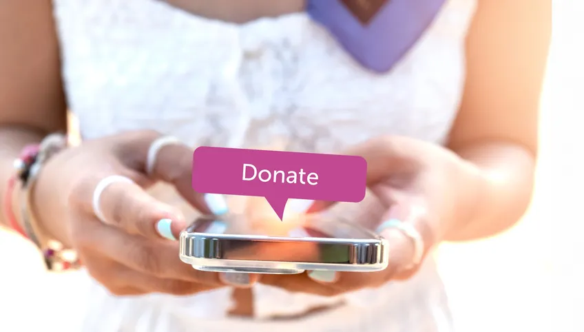 a person wearing a white dress and several bracelets types on a phone. A pop up bubble saying donate is displayed above the phone. 