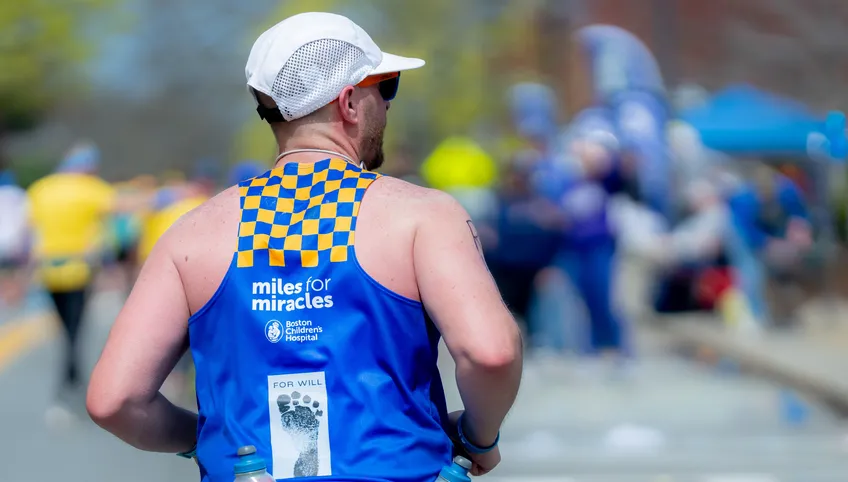 The back of a person wearing a customized blue and yellow boston children's hospital singlet. They are wearing a white hat and running on a busy marathon course