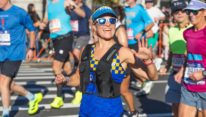 A smiling runner wearing sunglasses, a blue visor, and a hydration vest waves while running in a crowded road race, surrounded by other runners in colorful athletic gear.