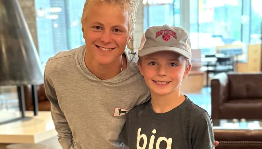 Two smiling boys pose together indoors; the older teen in a gray hoodie leans in with an arm around a younger boy wearing a gray cap and a dark T-shirt, with seating blurred in the background.
