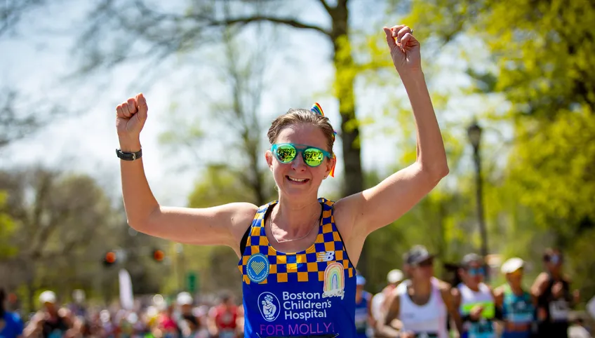 A runner wearing a blue and yellow Boston Children's singlet raises her arms triumphantly and smiles at the camera. l 