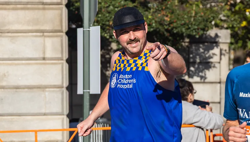 A person with a mustache smiles and points at the camera. He is wearing a blue and yellow boston children's racing singlet and a black hat. 