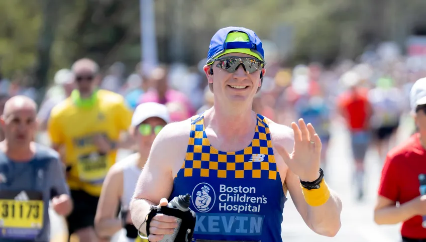 man holding water bottle waving at camera smiling while running a marathon