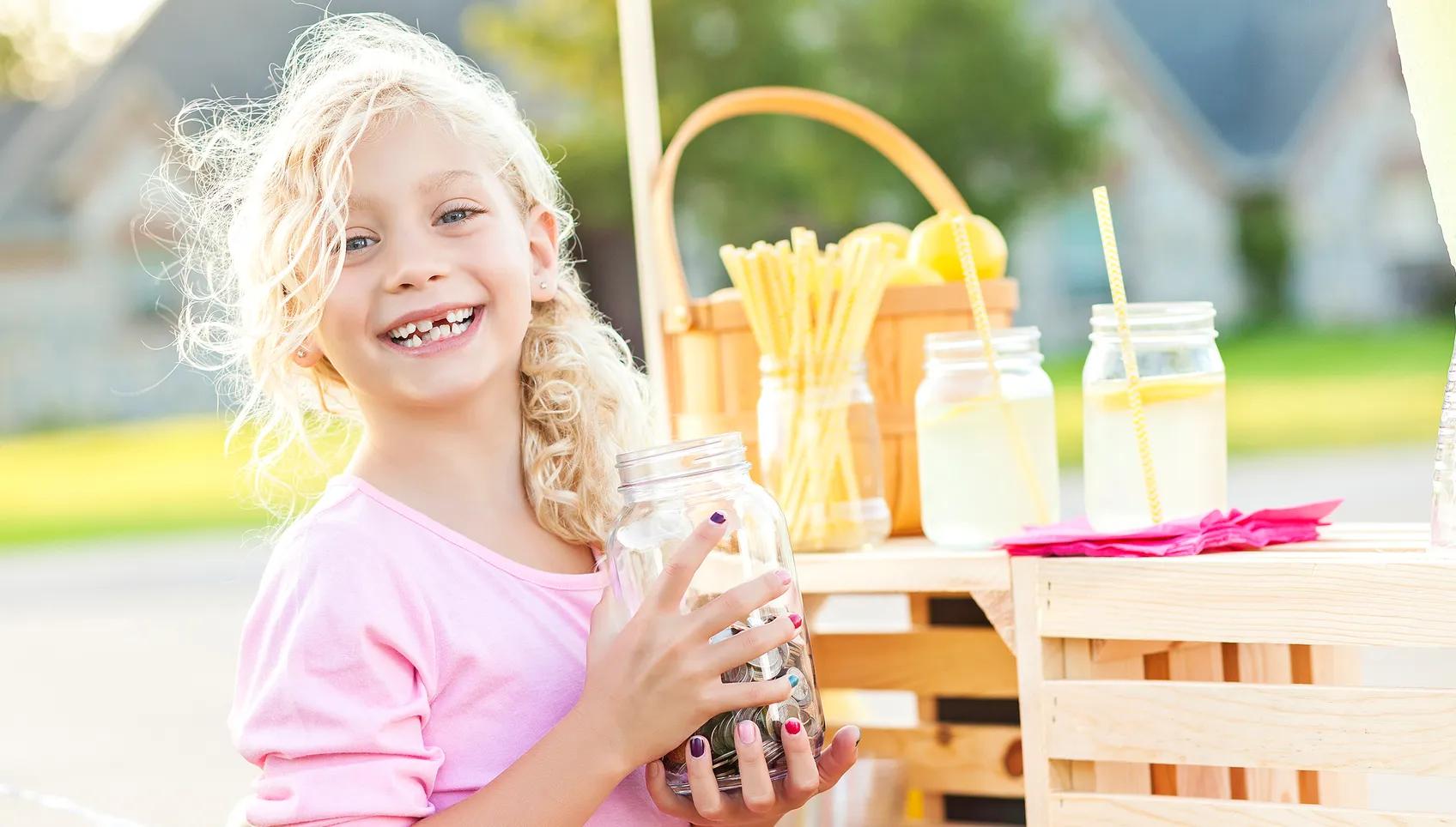 little girl holding a bottle of coins behind lemonade stand
