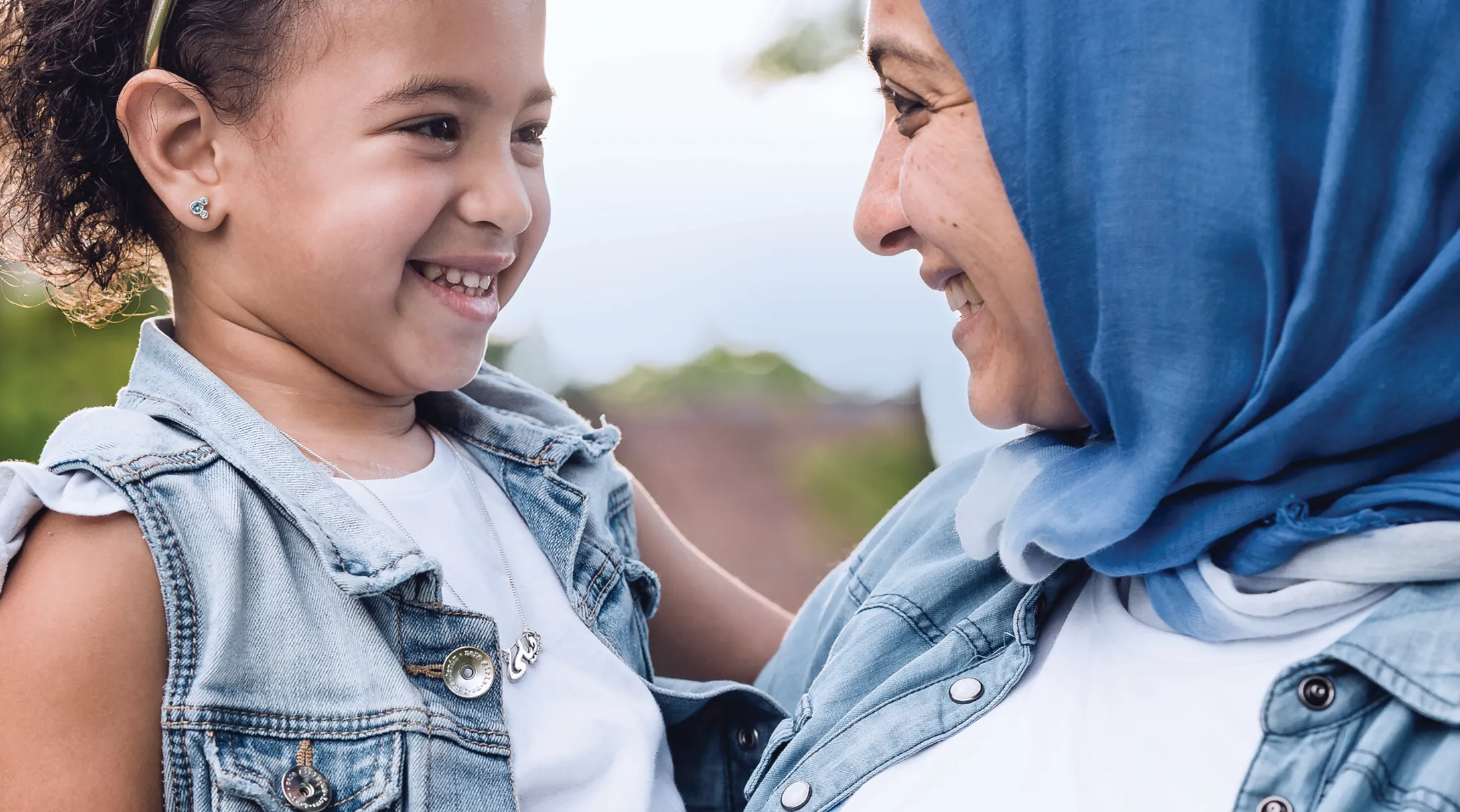 A young girl smiles at her mother. They wear matching clothes, and the mother wears a headscarf.
