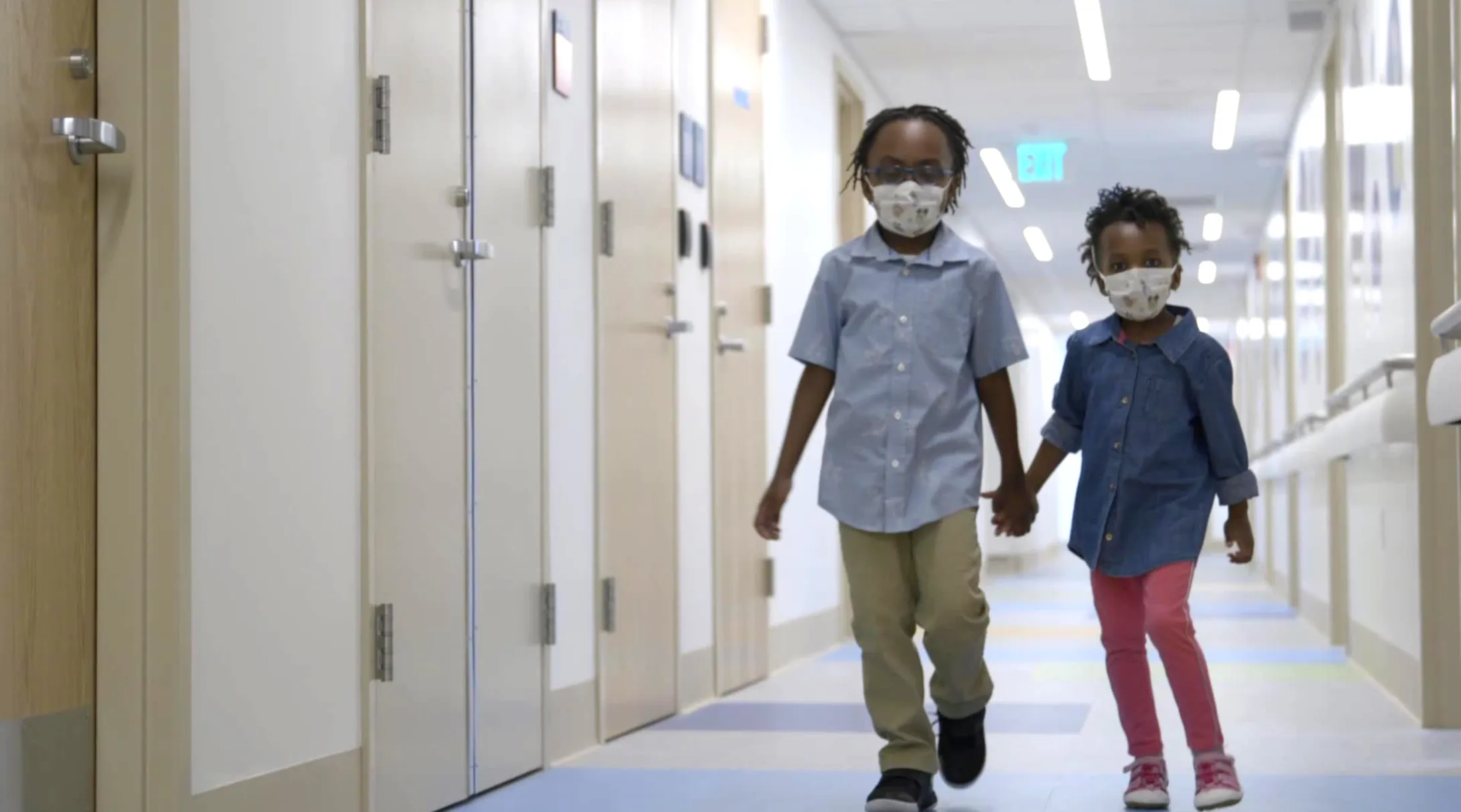 Two children wearing masks walk down a hallway holding hands