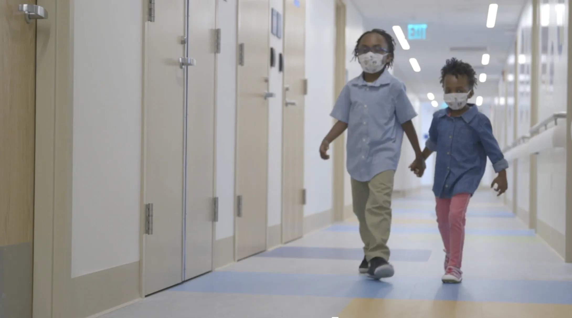 Two children with masks walking hand in hand together down hallway