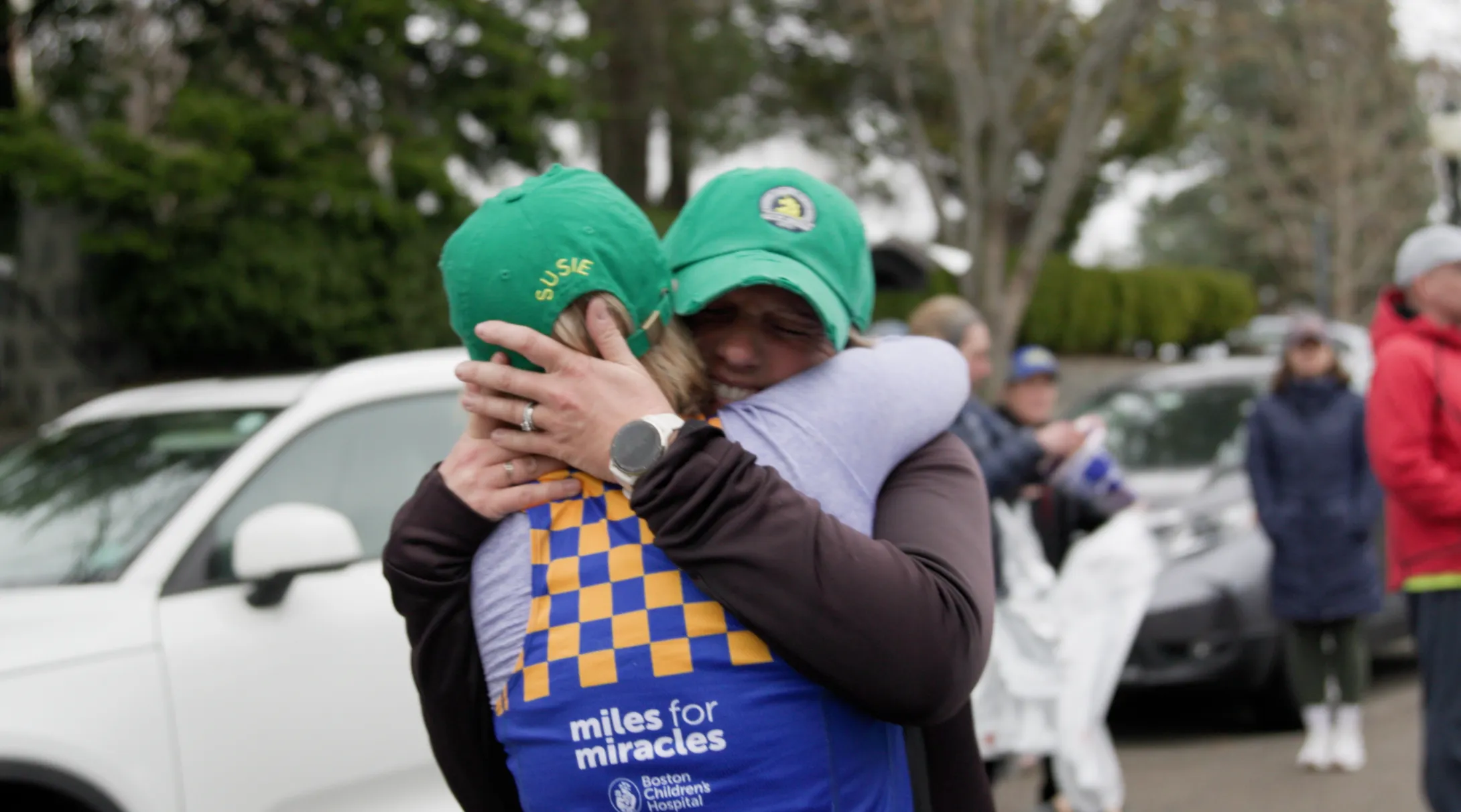 two people wearing bright green hats embrace. One person with their back to the camera wears a blue Boston Children's Hospital running singlet with yellow checks.  