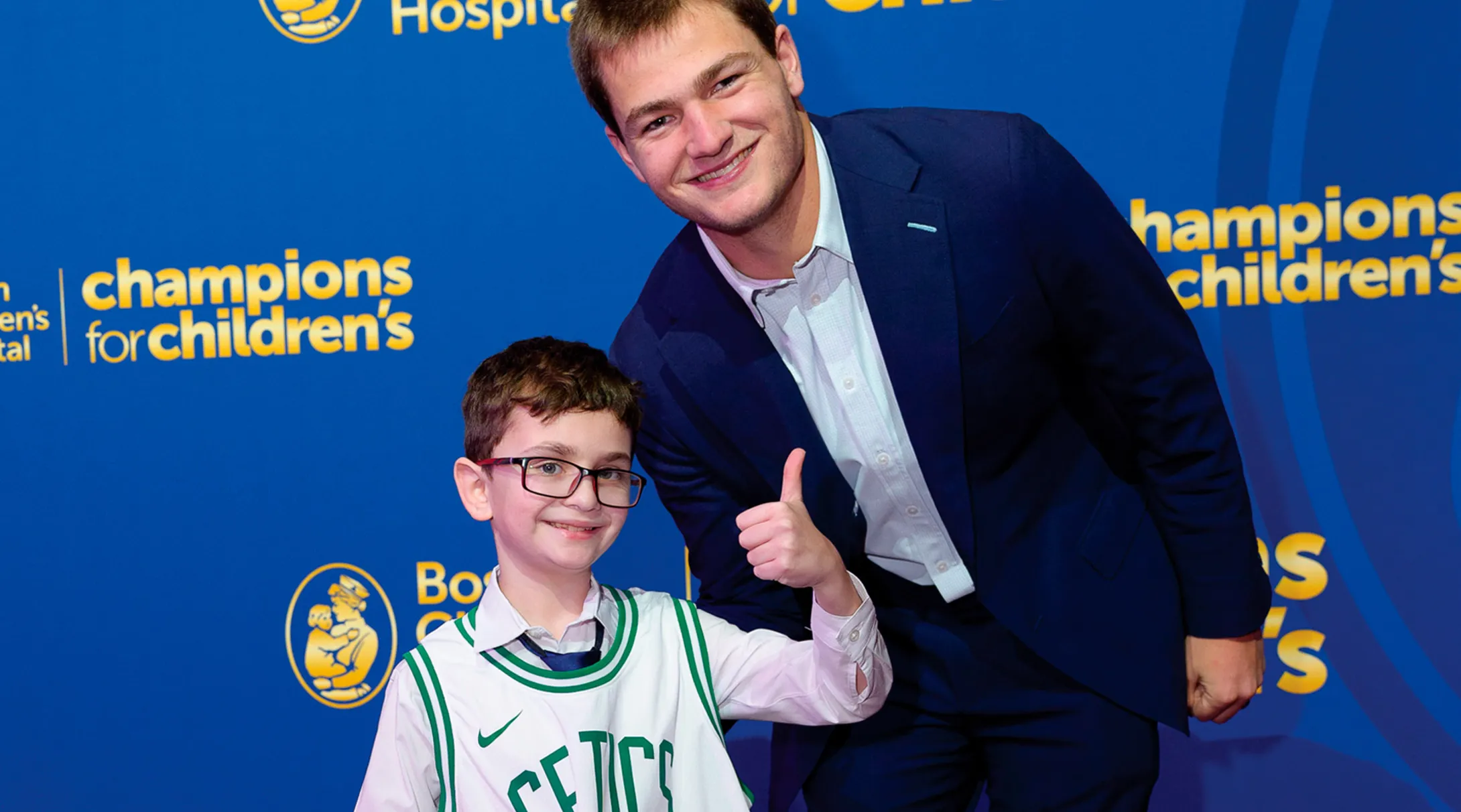 A smiling man in a navy suit leans in beside a smiling boy wearing glasses and a Celtics jersey; they pose in front of a blue “Champions for Children’s” Boston Children’s Hospital backdrop, with the boy giving a thumbs-up.