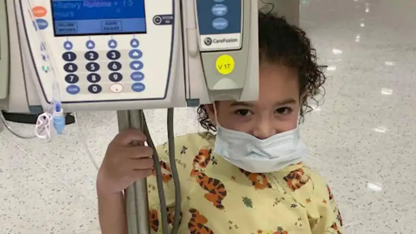 A young girl with curly hair stands behind a hospital monitor. She is wearing a mask, but you can still see a mischievous smile in her eyes.