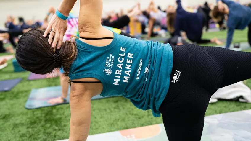 A woman wearing a Miracle Makers tank top reaches into a side plank on her yoga mat