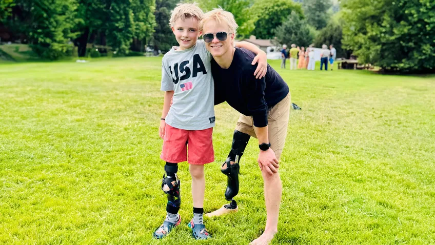 An adult wearing sunglasses and a leg prosthesis kneels on a grassy field with an arm around a smiling child in a “USA” T-shirt, red shorts, and a similar leg prosthesis, with trees and a small group of people in the background.