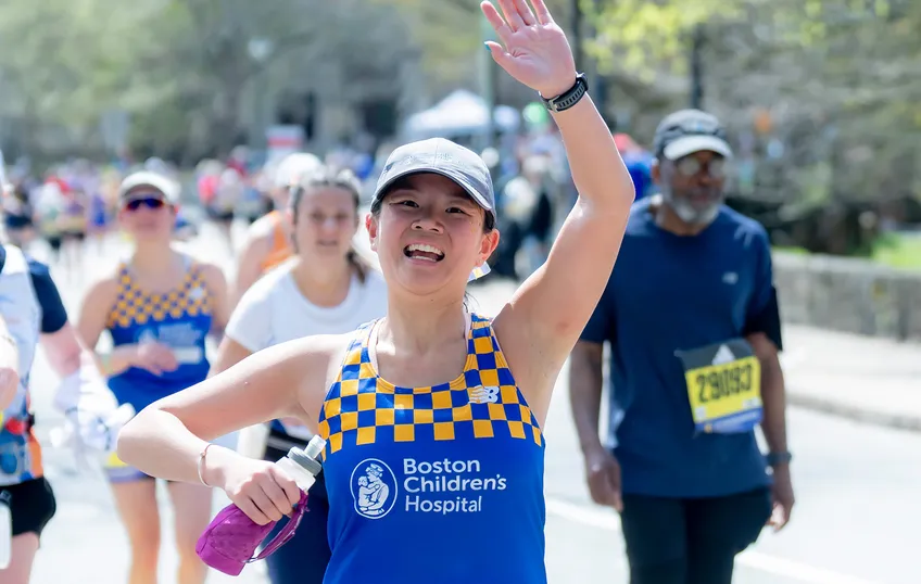 A person wearing a navy bule baseball cap and bright blue and yellow check Boston Children's Hospital singlet runs with her arm raised waving.