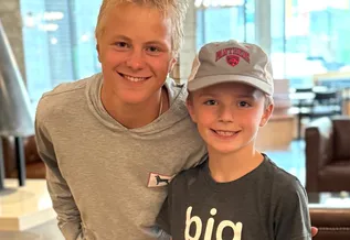 Two smiling boys pose together indoors; the older teen in a gray hoodie leans in with an arm around a younger boy wearing a gray cap and a dark T-shirt, with seating blurred in the background.