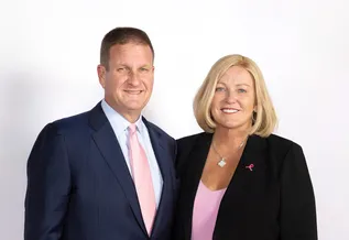 Rob Hale (a man with dark, close cropped hair wearing a suit and a pink tie) stands with his wife, Karen Hale ( a woman with shoulder length blonde hair wearing a dark jacket and a pink shirt). The two are smiling.